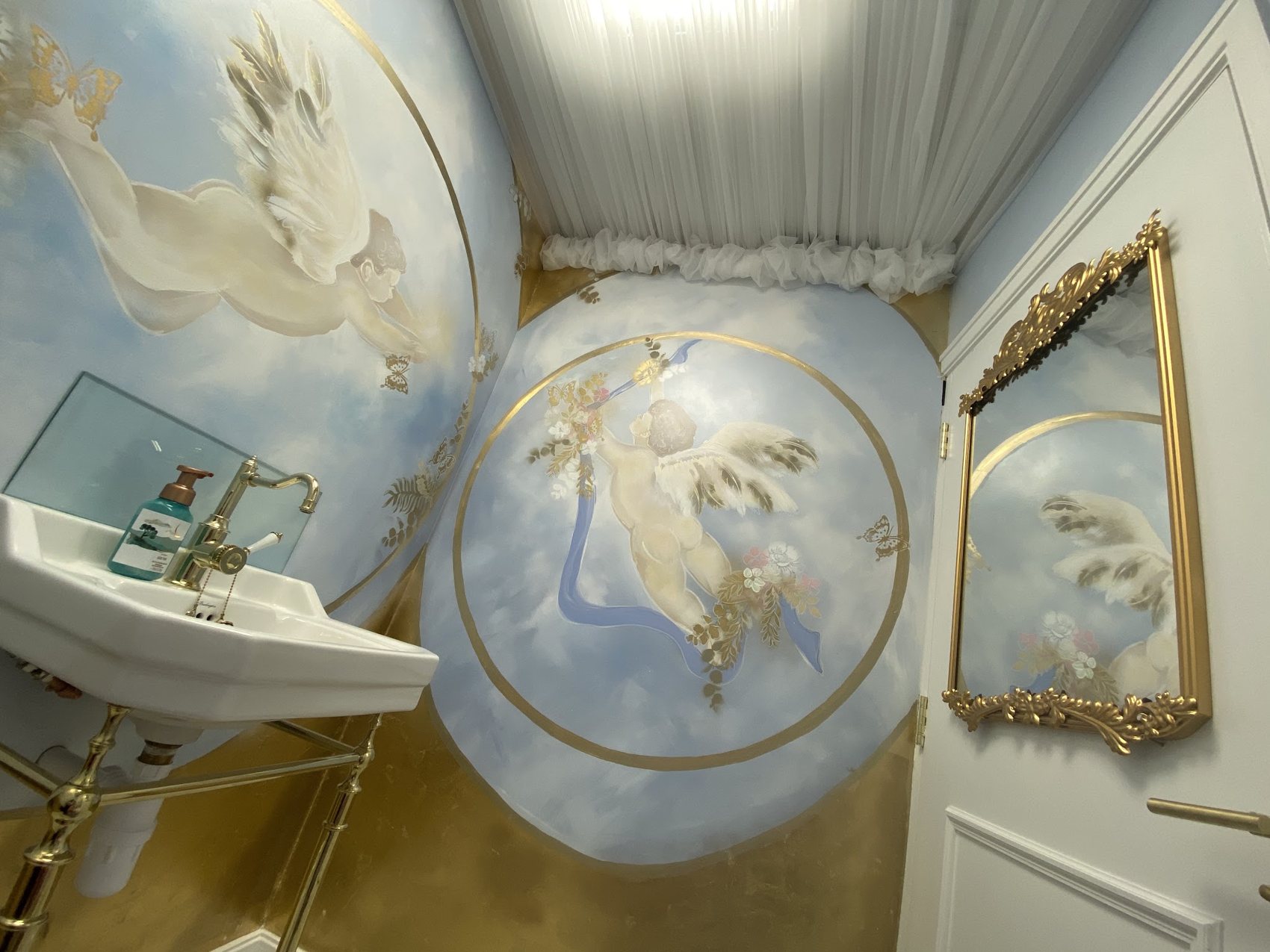 A washroom painted with angels in gold and sky blue, with a gold-framed mirror