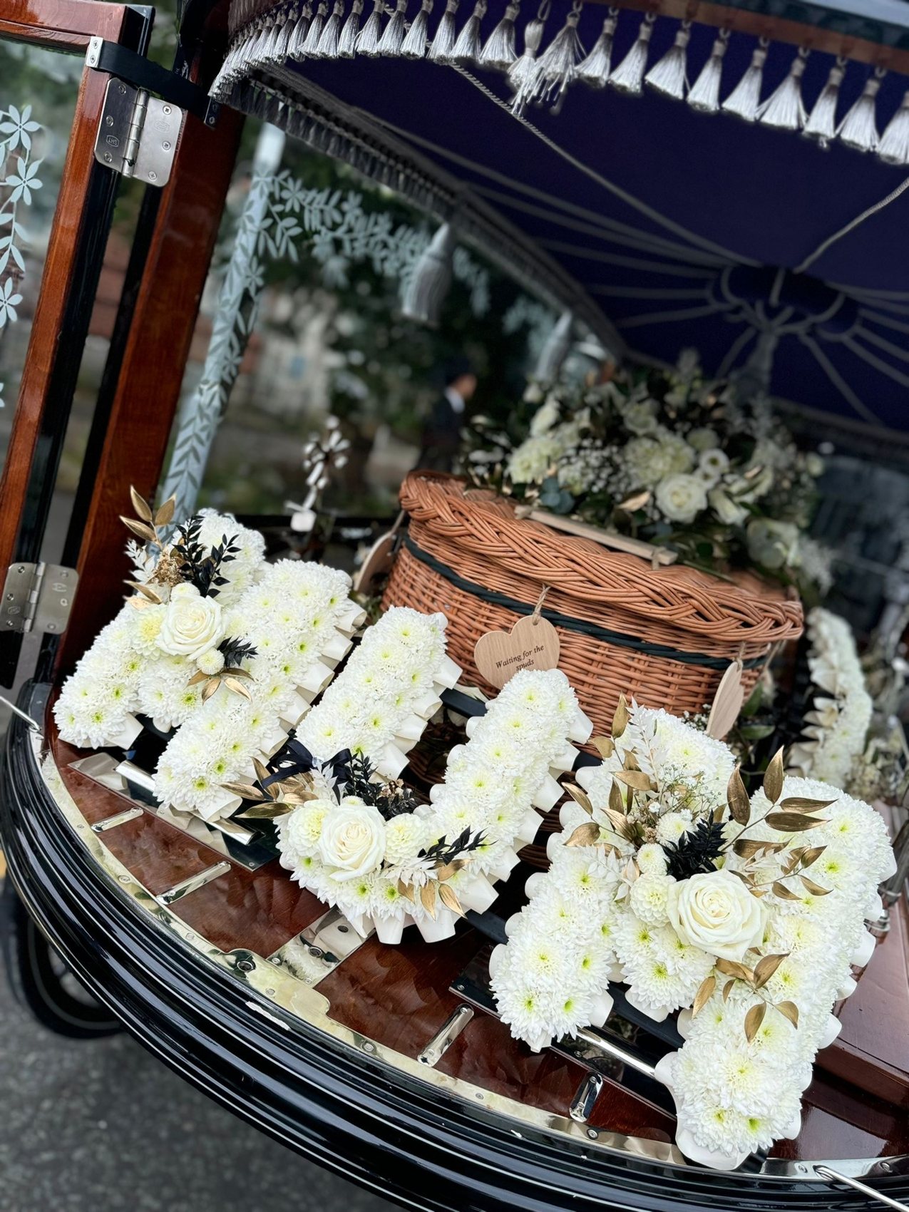 A glass-sided horse-drawn hearse carrying a white floral tribute spelling MUM and a wicker casket